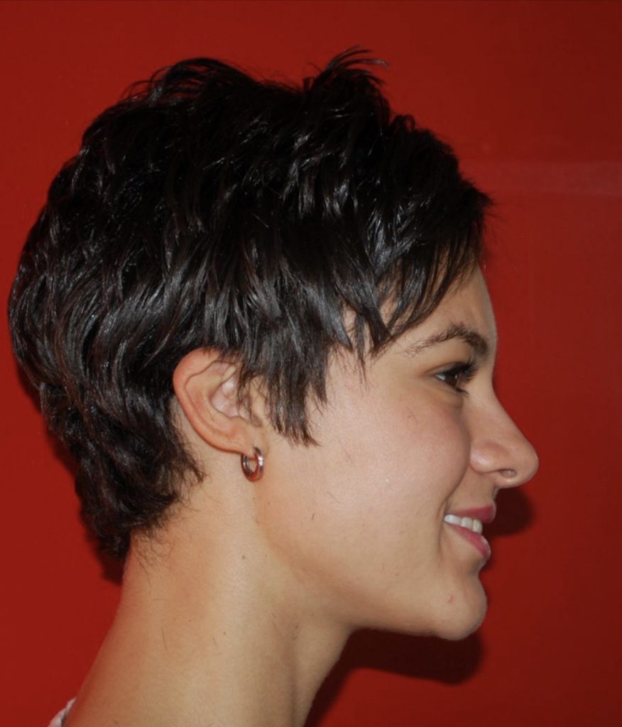 Modern textured brunette pixie haircut with soft layers, side fringe, and tapered neckline photographed in profile against a red background at Salon International in Mount Pleasant, SC.